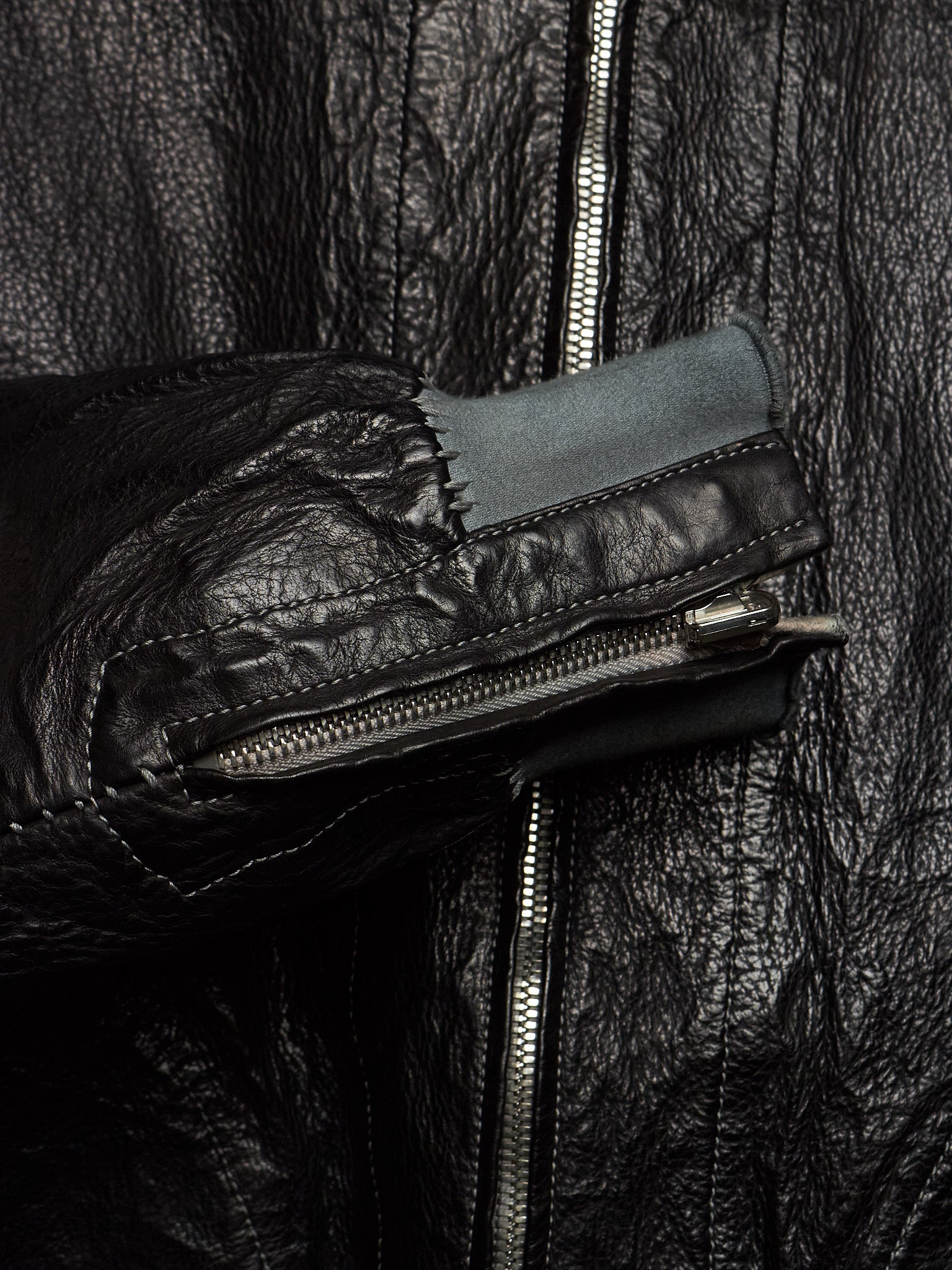 Close-up of a black leather jacket with a prominent front zipper, a zippered pocket on the sleeve, and a contrasting dark gray fabric lining.