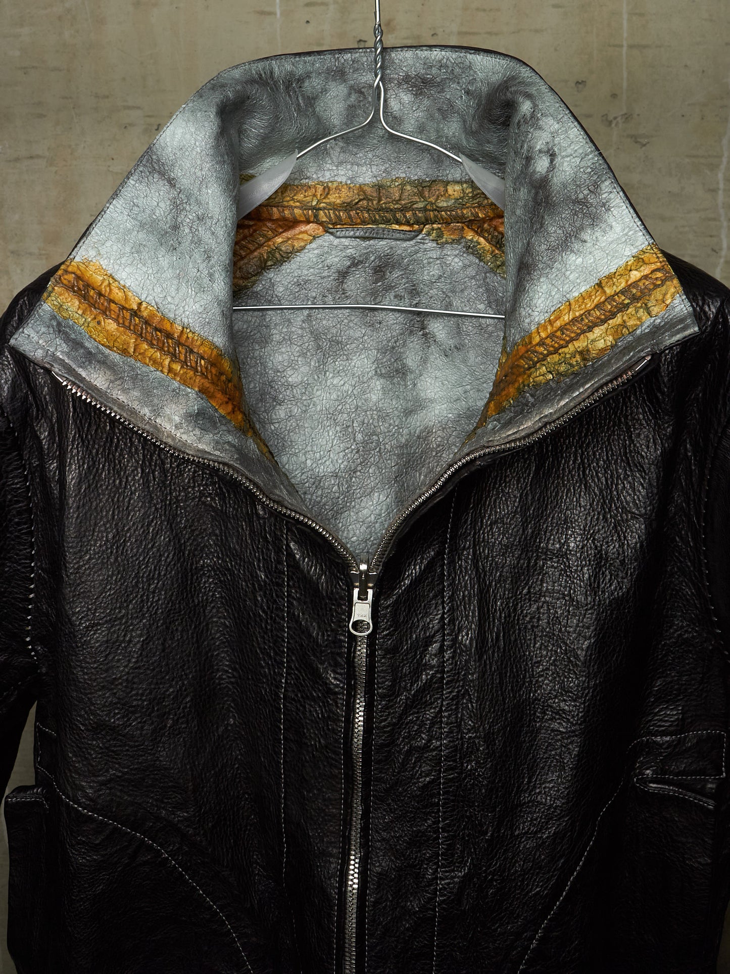 A black leather jacket with a high collar, featuring a gray faux-fur lining and a distinctive gold and brown patterned inner band, shown hanging against a concrete wall.