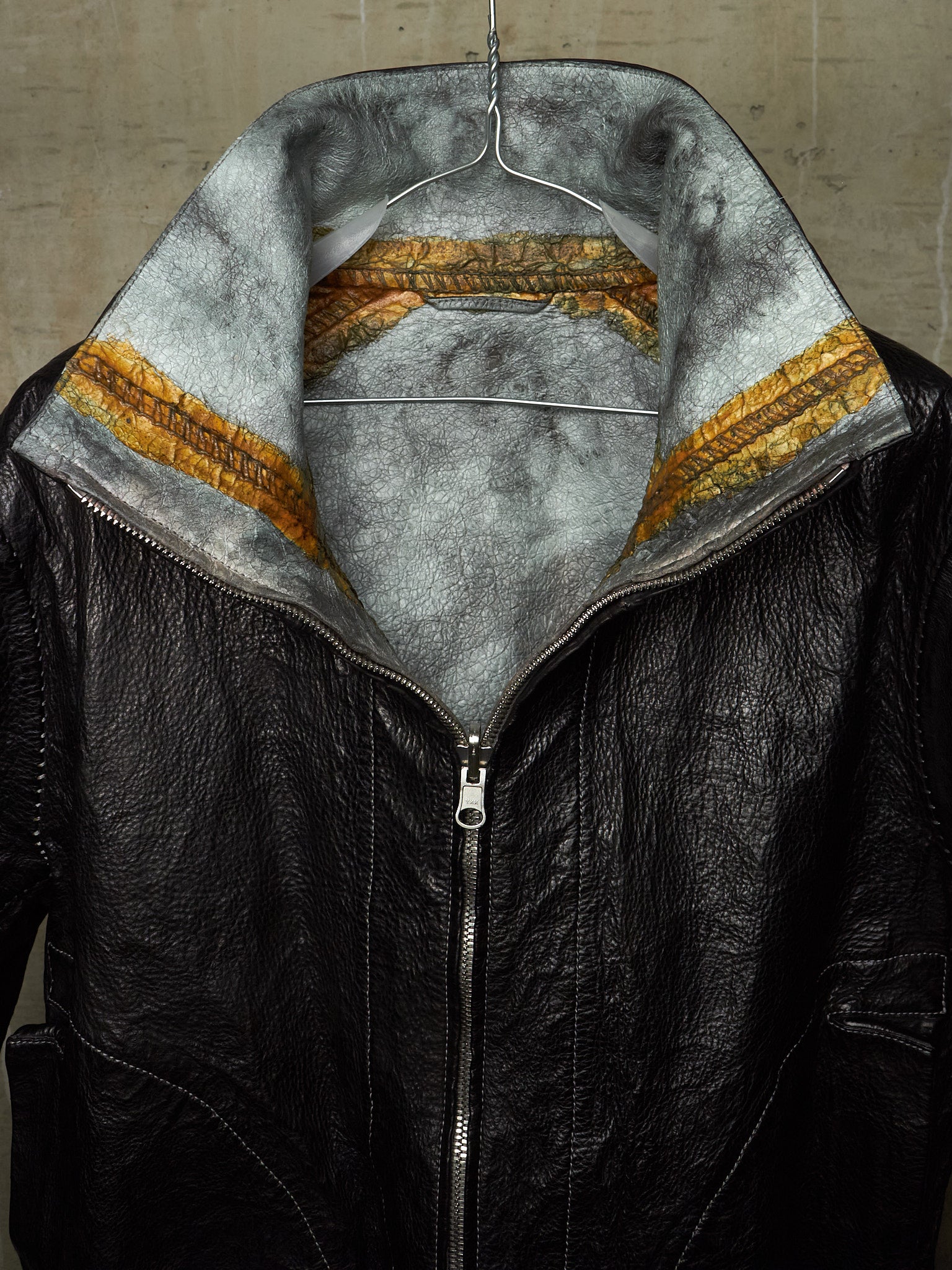 A black leather jacket with a high collar, featuring a gray faux-fur lining and a distinctive gold and brown patterned inner band, shown hanging against a concrete wall.