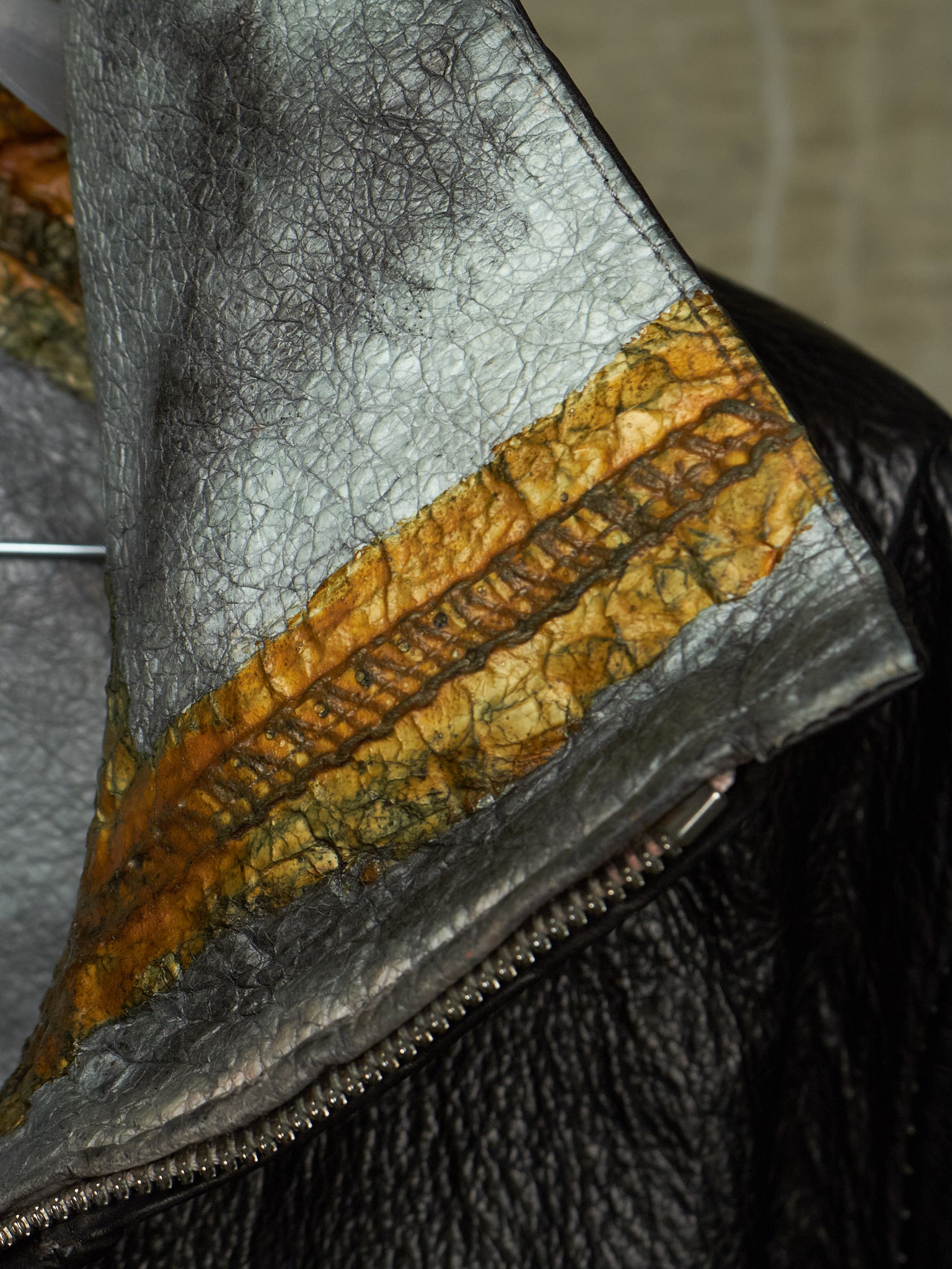 Close-up of a leather jacket collar showing a textured silver-gray leather body, a wide mustard-yellow and brown patterned band, and a black leather section with a metal zipper.