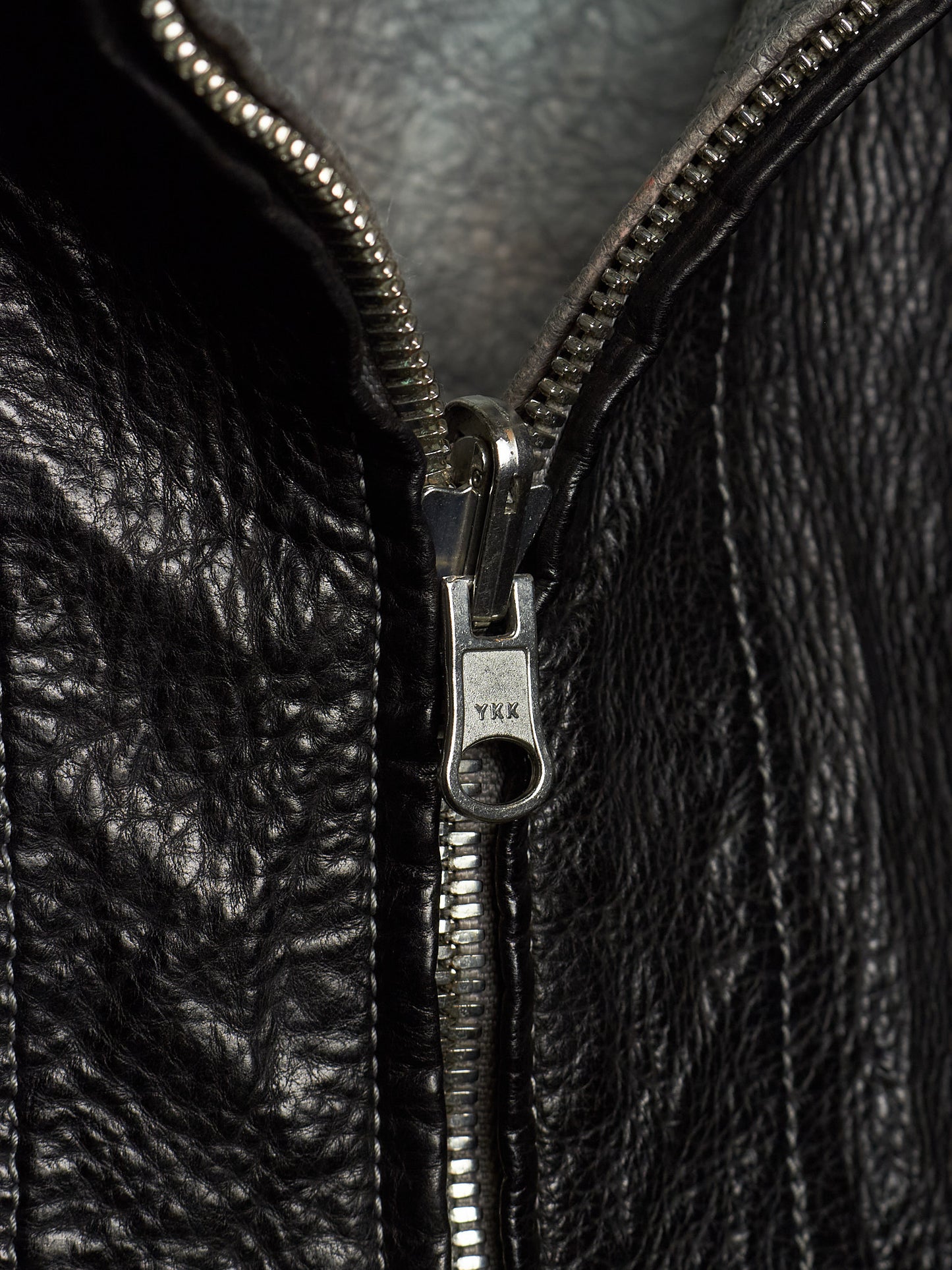 Close-up of a black leather jacket with a silver metal zipper and pull tab, showing the texture of the leather and the engraved brand name YKK.