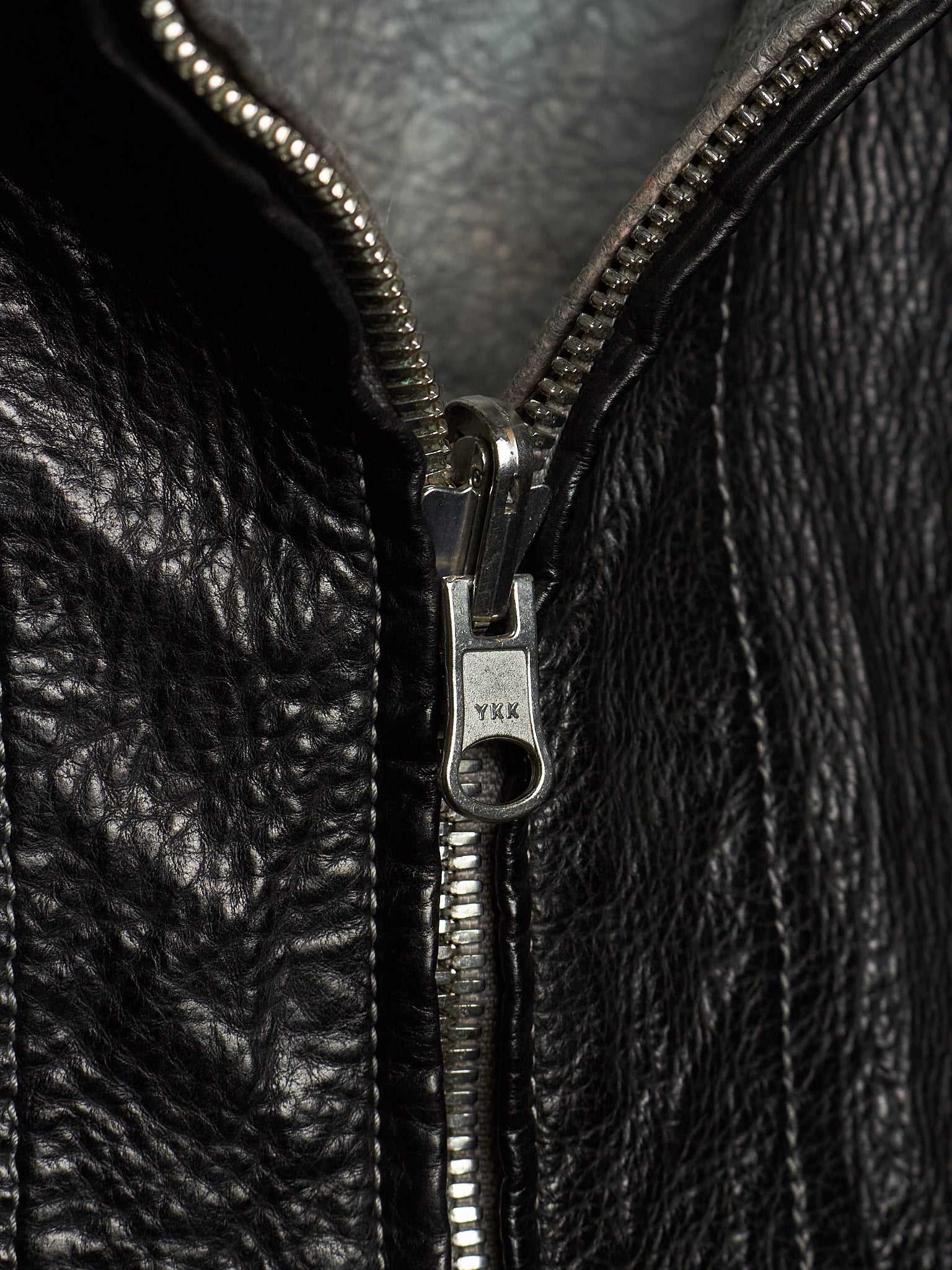 Close-up of a black leather jacket with a silver metal zipper and pull tab, showing the texture of the leather and the engraved brand name YKK.