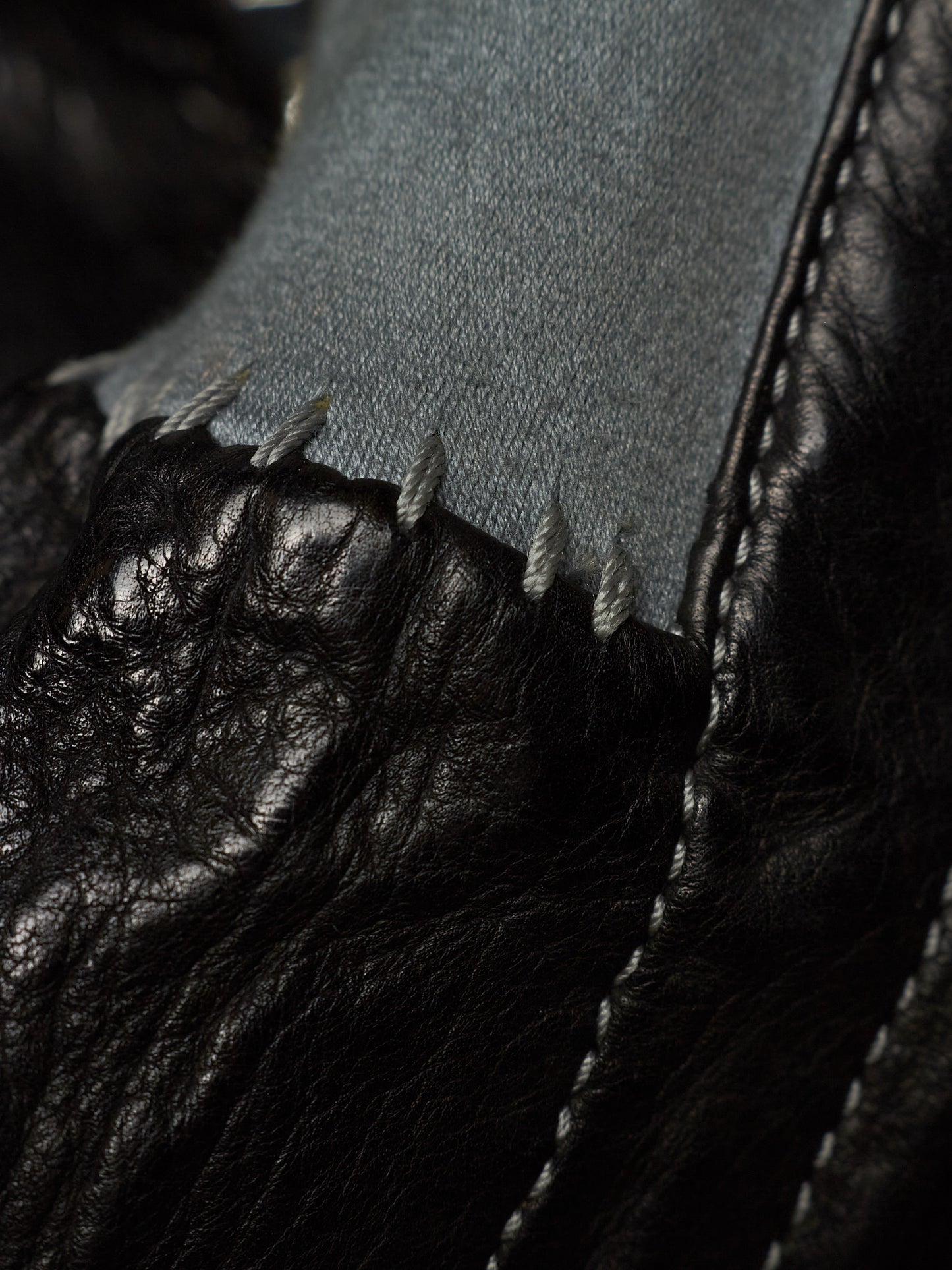 Close-up detail of a black leather shoe with a raw, frayed edge and white stitching, showing a light gray fabric lining.