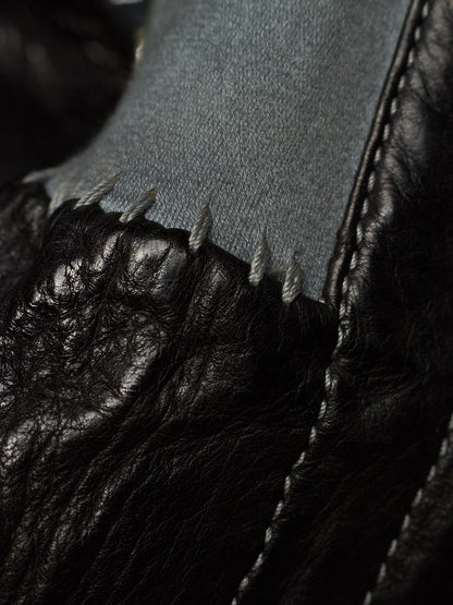 Close-up detail of a black leather shoe with a raw, frayed edge and white stitching, showing a light gray fabric lining.
