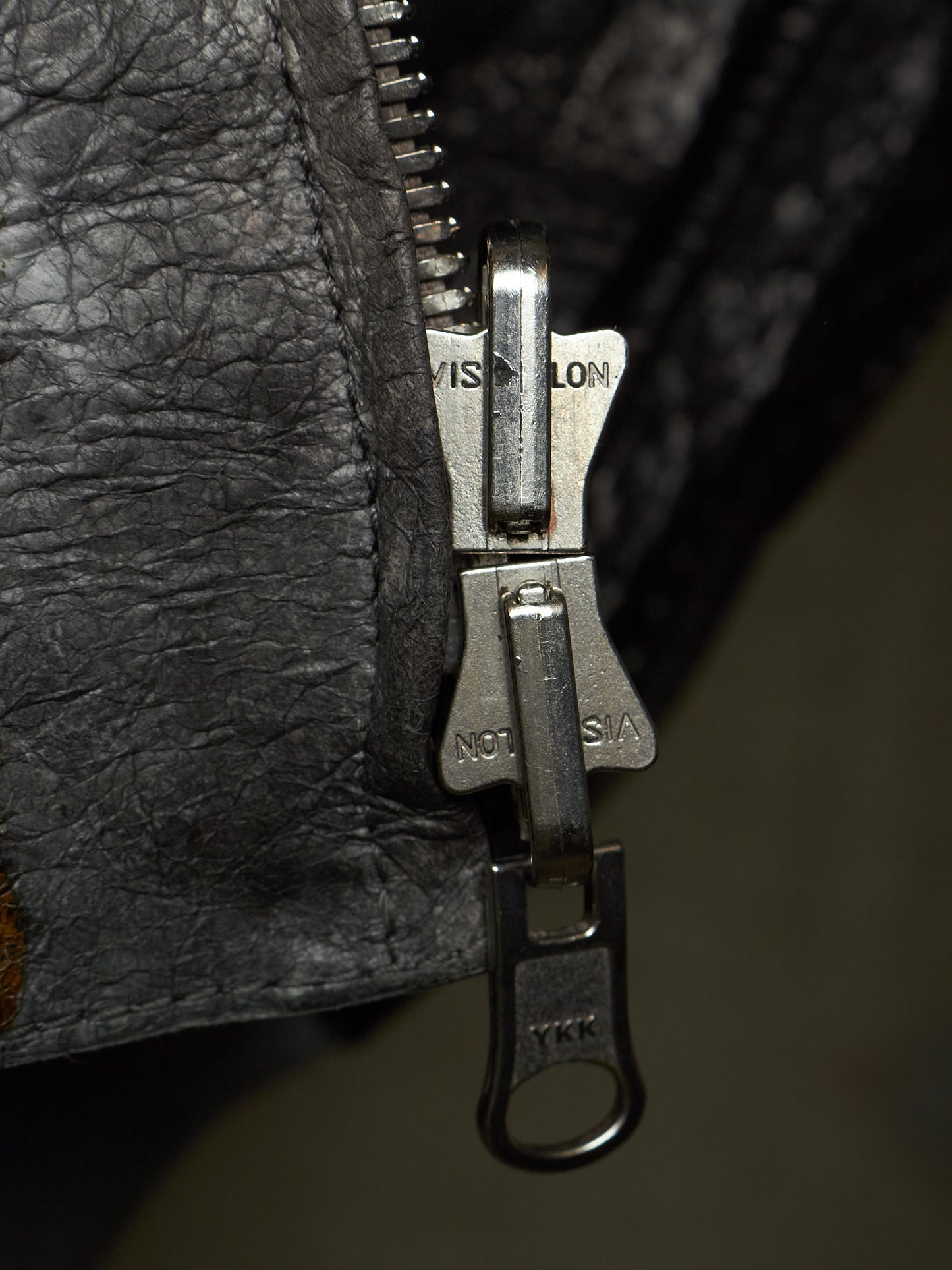 Close-up of a silver metal zipper pull with 'VISALON' and 'NOT A COPY' branding, set against a dark textured black leather background.