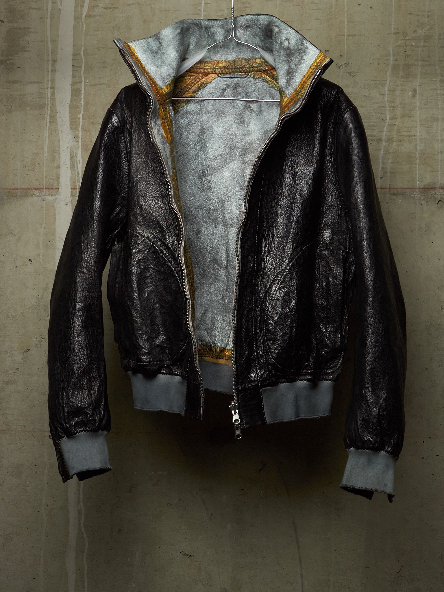 A black leather bomber jacket with a gray interior lining and gold patterned trim along the collar, hanging against a textured concrete wall.