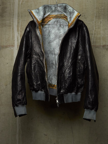 A black leather bomber jacket with a gray interior lining and gold patterned trim along the collar, hanging against a textured concrete wall.