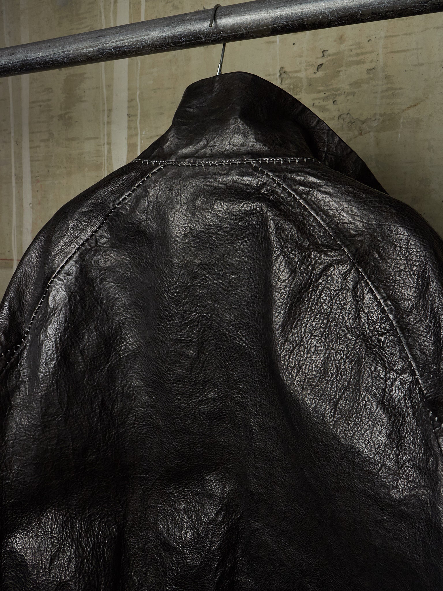 Close-up of a black leather jacket hanging on a metal clothing rack against a textured beige wall.