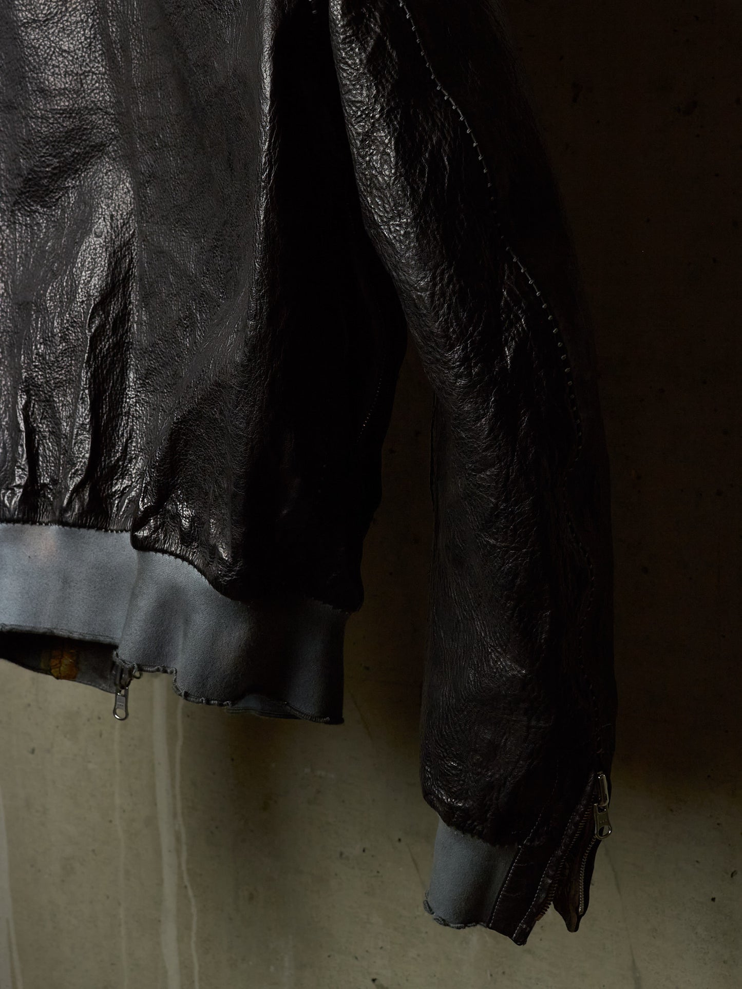 Close-up of a black leather jacket with a raw, frayed hem and cuffs, hanging against a textured concrete wall.