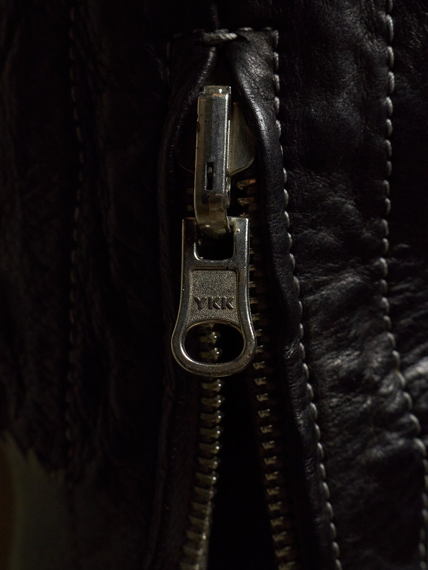 Close-up of a black leather jacket featuring a silver metal zipper with a pull tab engraved with the YKK logo.