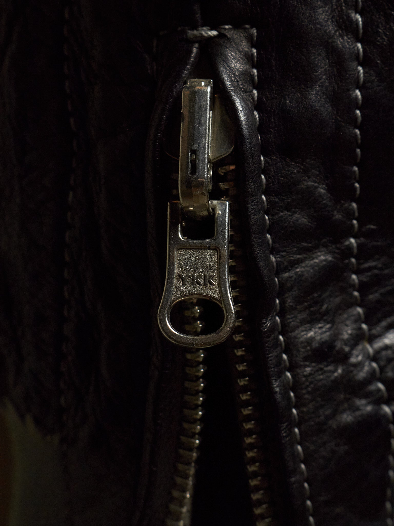 Close-up of a black leather jacket featuring a silver metal zipper with a pull tab engraved with the YKK logo.