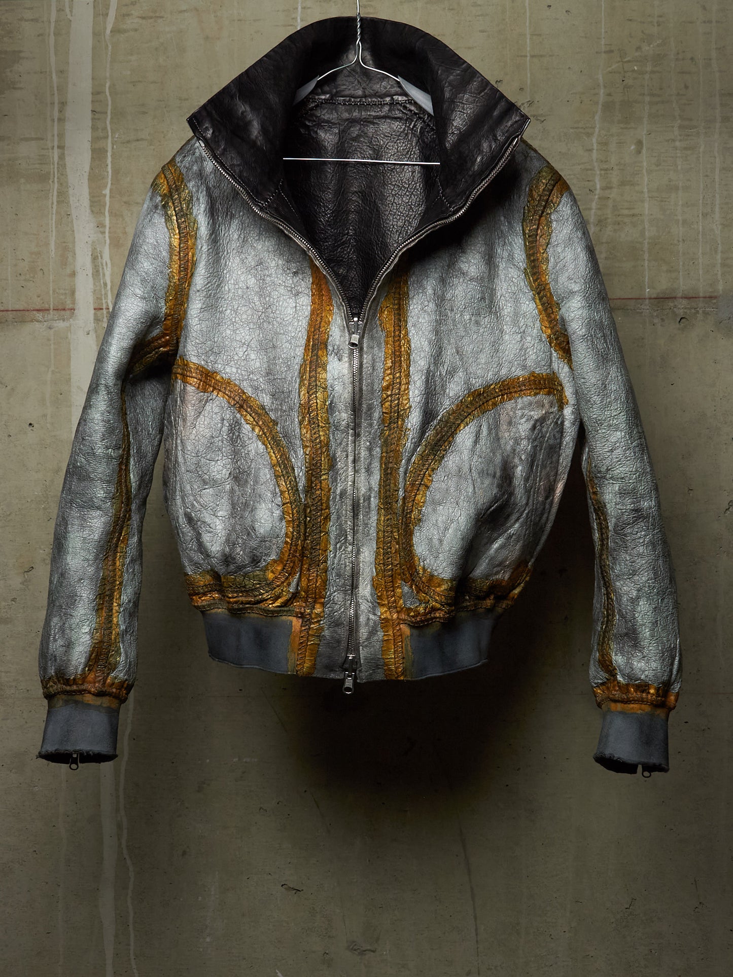 A distressed metallic silver leather jacket with gold zippers and trim, hanging against a textured concrete wall.