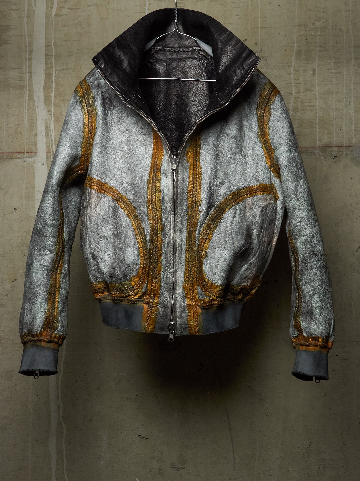 A distressed metallic silver leather jacket with gold zippers and trim, hanging against a textured concrete wall.