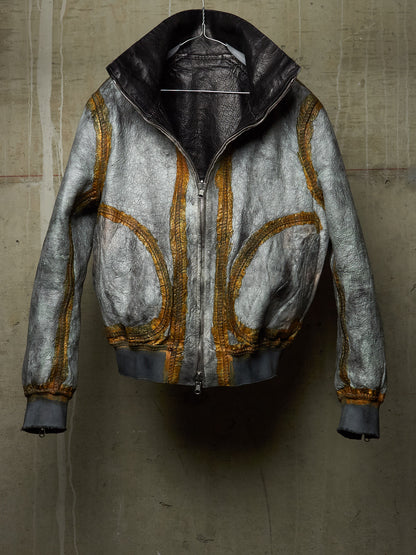 A distressed metallic silver leather jacket with gold zippers and trim, hanging against a textured concrete wall.