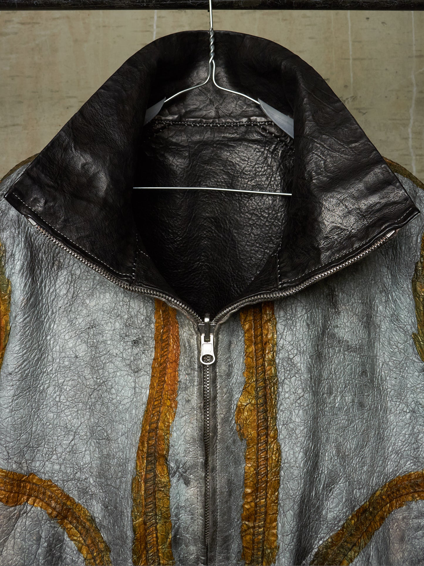 A close-up of a textured leather jacket hanging on a wire hanger against a neutral background. The jacket features a distressed silver-gray body with vertical gold-colored stitched stripes and a contrasting black leather collar.