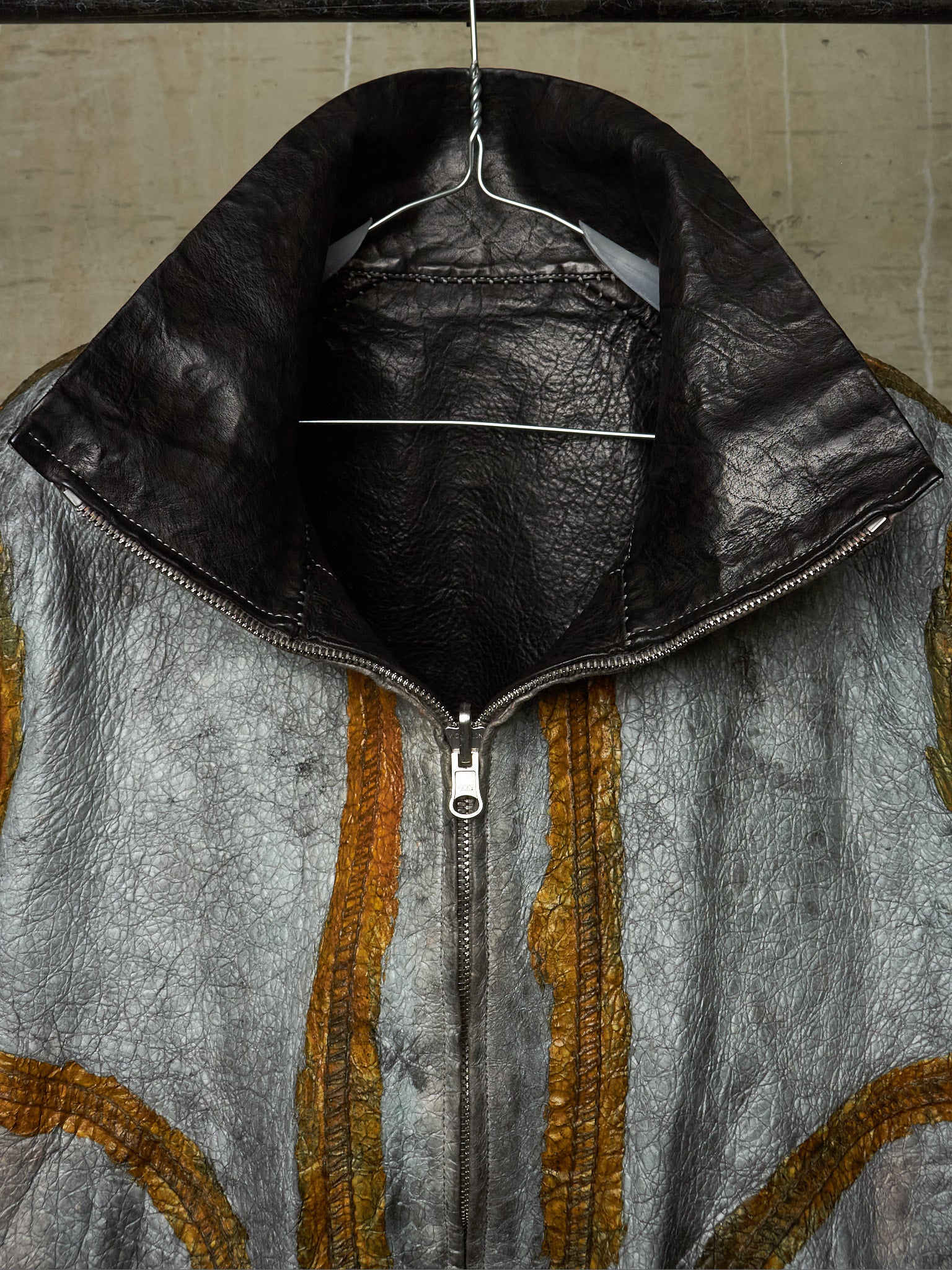 A close-up of a textured leather jacket hanging on a wire hanger against a neutral background. The jacket features a distressed silver-gray body with vertical gold-colored stitched stripes and a contrasting black leather collar.