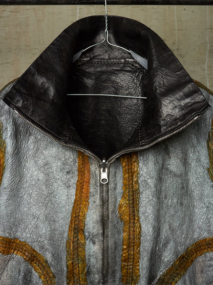 A close-up of a textured leather jacket hanging on a wire hanger against a neutral background. The jacket features a distressed silver-gray body with vertical gold-colored stitched stripes and a contrasting black leather collar.