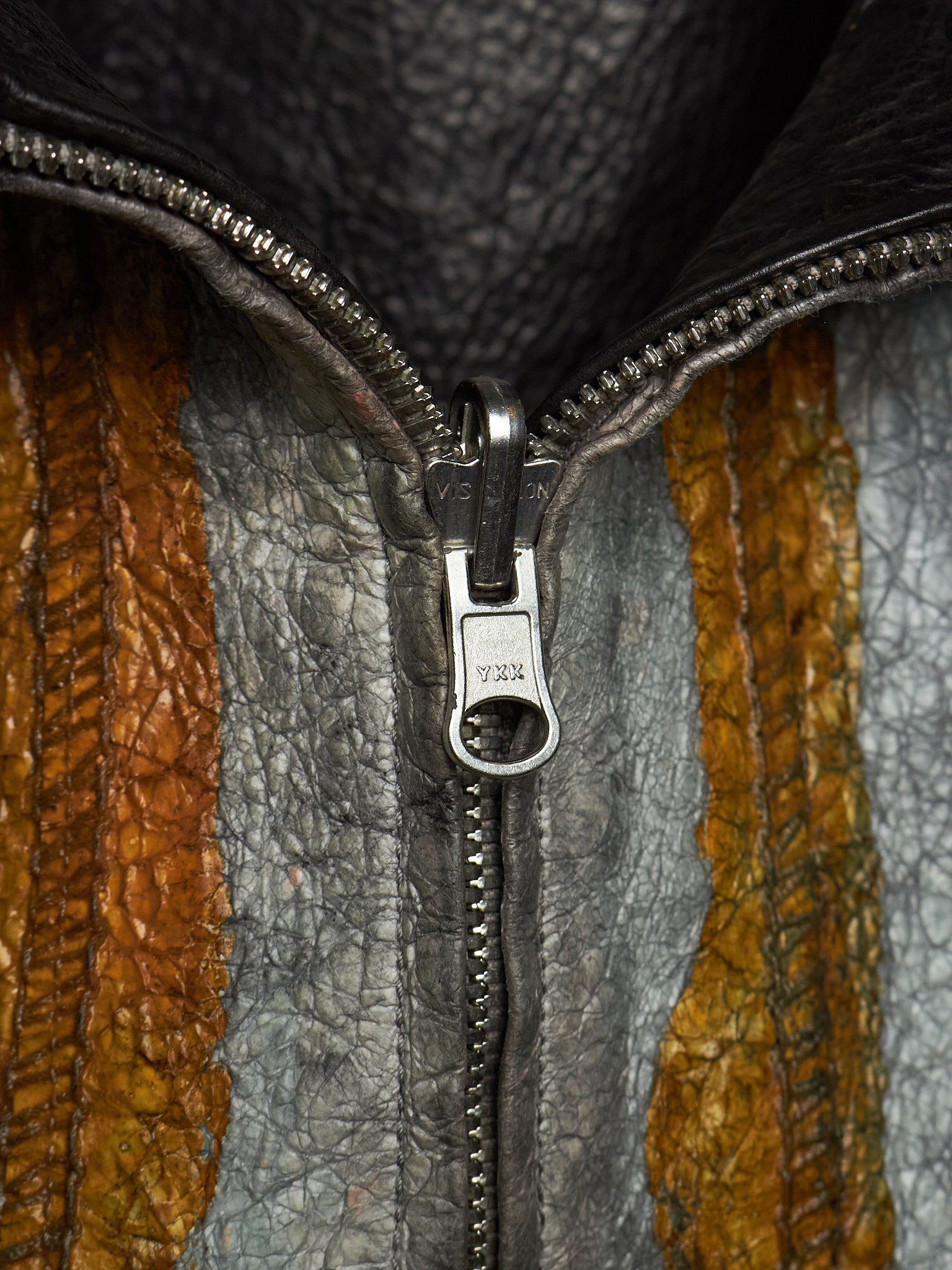Close-up of a textured leather jacket with a silver metal zipper and pull tab, showing vertical panels of gray, brown, and mustard yellow leather.