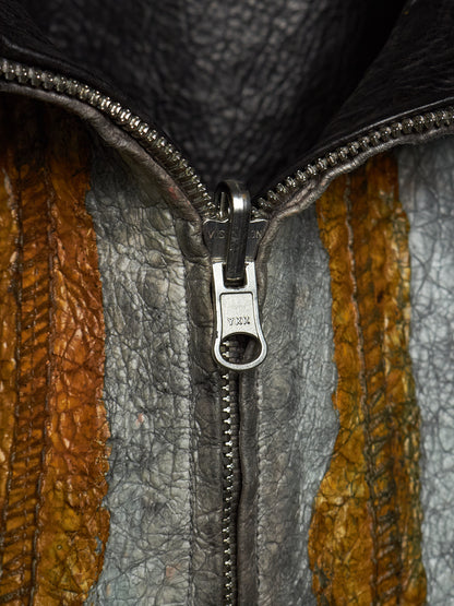 Close-up of a textured leather jacket with a silver metal zipper and pull tab, showing vertical panels of gray, brown, and mustard yellow leather.