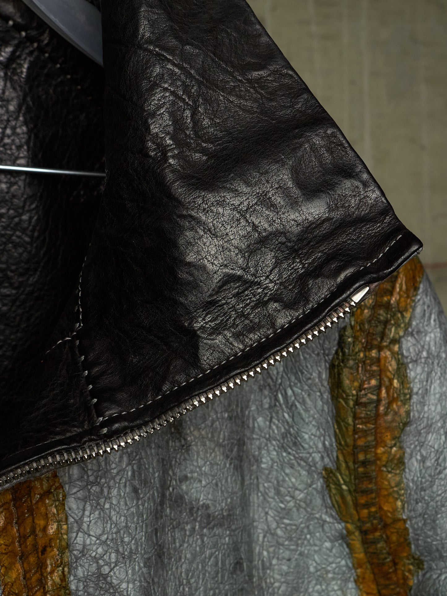 Close-up detail of a black leather jacket with a silver zipper, showing the texture of the leather and a portion of a gray and orange patterned garment underneath.