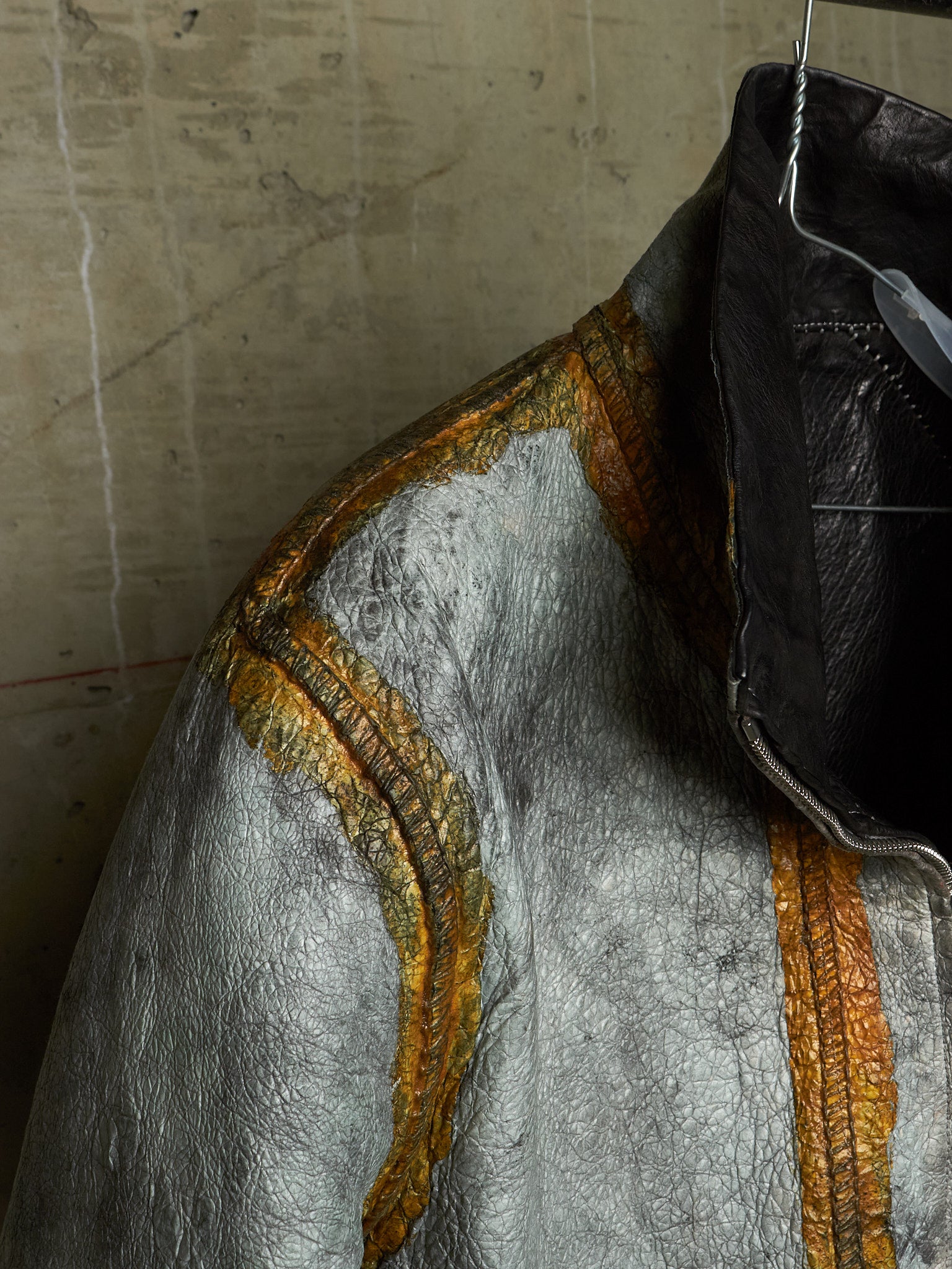 A close-up of a textured silver jacket with metallic gold-orange stitched detailing, hanging against a concrete wall.