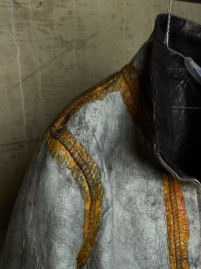 A close-up of a textured silver jacket with metallic gold-orange stitched detailing, hanging against a concrete wall.