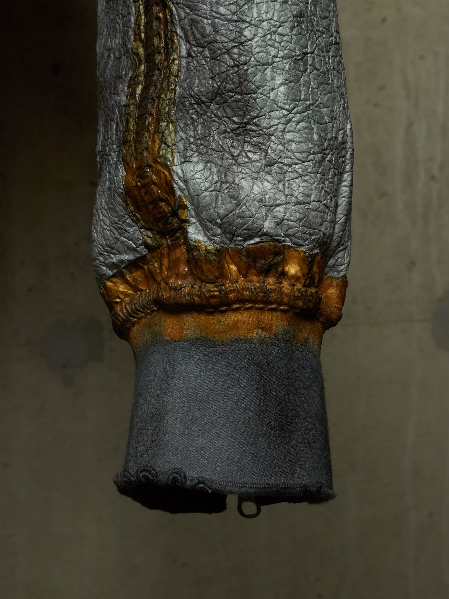 A close-up of a textured silver leather or faux-leather sleeve with a distressed gold braided cuff and a dark gray fabric cuff below, shown against a plain concrete-like background.