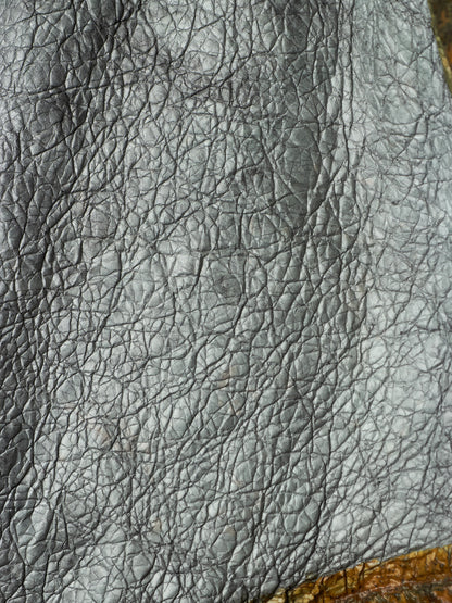 A close-up of dark gray pebbled leather texture with visible grain and a small warm brown area at the bottom right.
