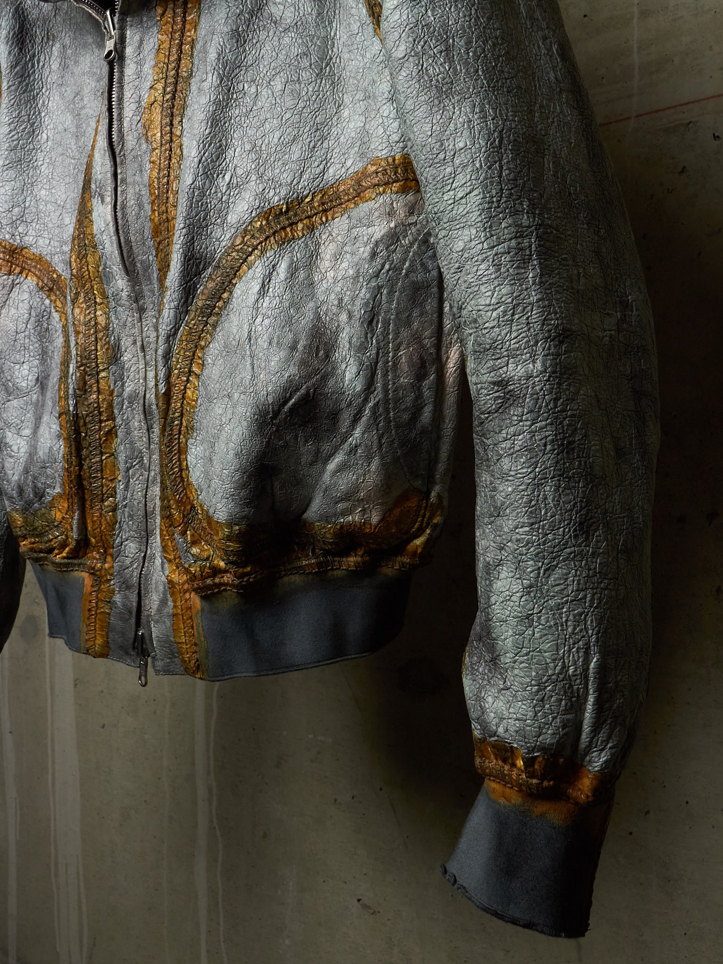 A close-up of a metallic silver jacket with gold embroidered detailing and dark gray ribbed cuffs and hem, shown hanging against a textured concrete wall.