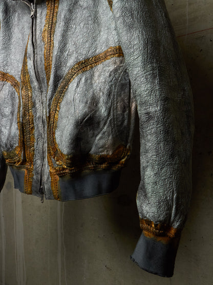 A close-up of a metallic silver jacket with gold embroidered detailing and dark gray ribbed cuffs and hem, shown hanging against a textured concrete wall.