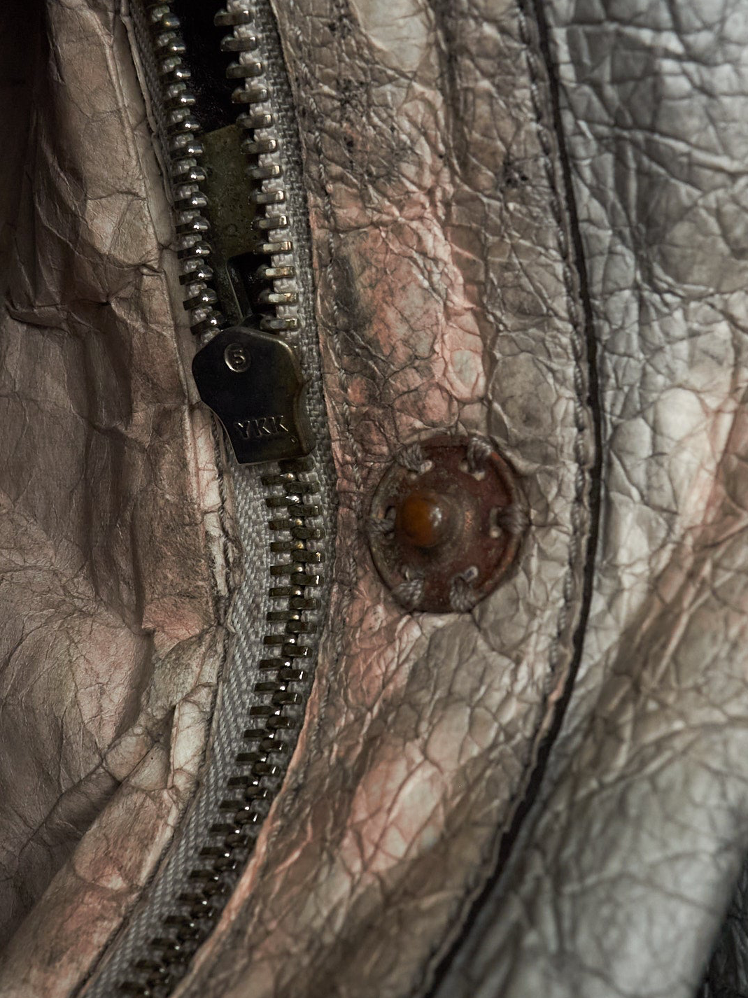 Extreme close-up of a distressed, heavily textured leather jacket featuring a silver-toned metal zipper with a dark pull tab engraved with 'YIK', a rusted metal rivet, and a heavily wrinkled, cracked leather surface in muted gray, brown, and copper tones.