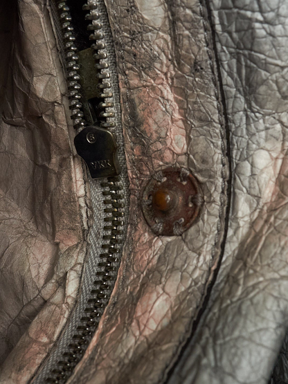 Extreme close-up of a distressed, heavily textured leather jacket featuring a silver-toned metal zipper with a dark pull tab engraved with 'YIK', a rusted metal rivet, and a heavily wrinkled, cracked leather surface in muted gray, brown, and copper tones.