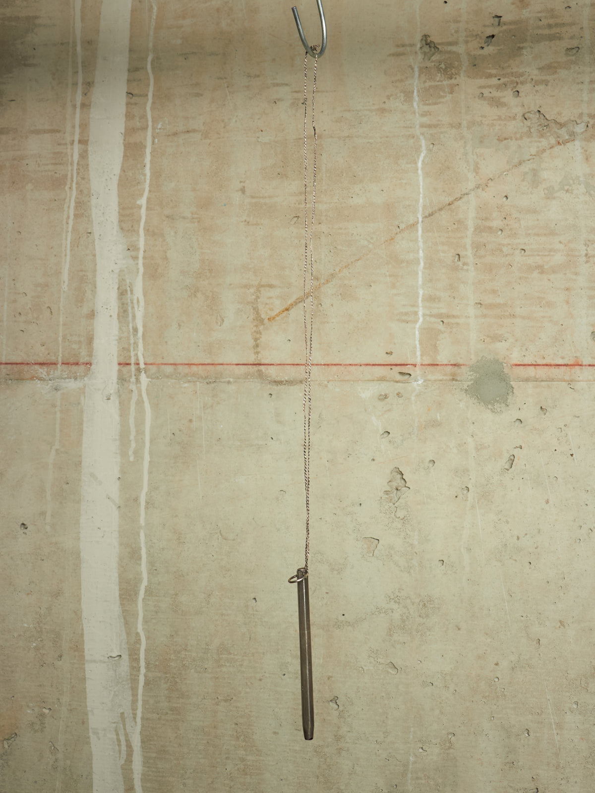 A minimalist art piece featuring a long, slender dark metal rod hanging from a chain from a metal hook on a textured concrete wall with vertical white paint drips and a thin red horizontal line.