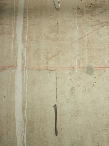 A minimalist art piece featuring a long, slender dark metal rod hanging from a chain from a metal hook on a textured concrete wall with vertical white paint drips and a thin red horizontal line.