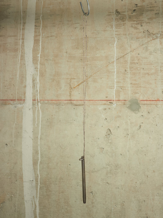 A minimalist art piece featuring a long, slender dark metal rod hanging from a chain from a metal hook on a textured concrete wall with vertical white paint drips and a thin red horizontal line.