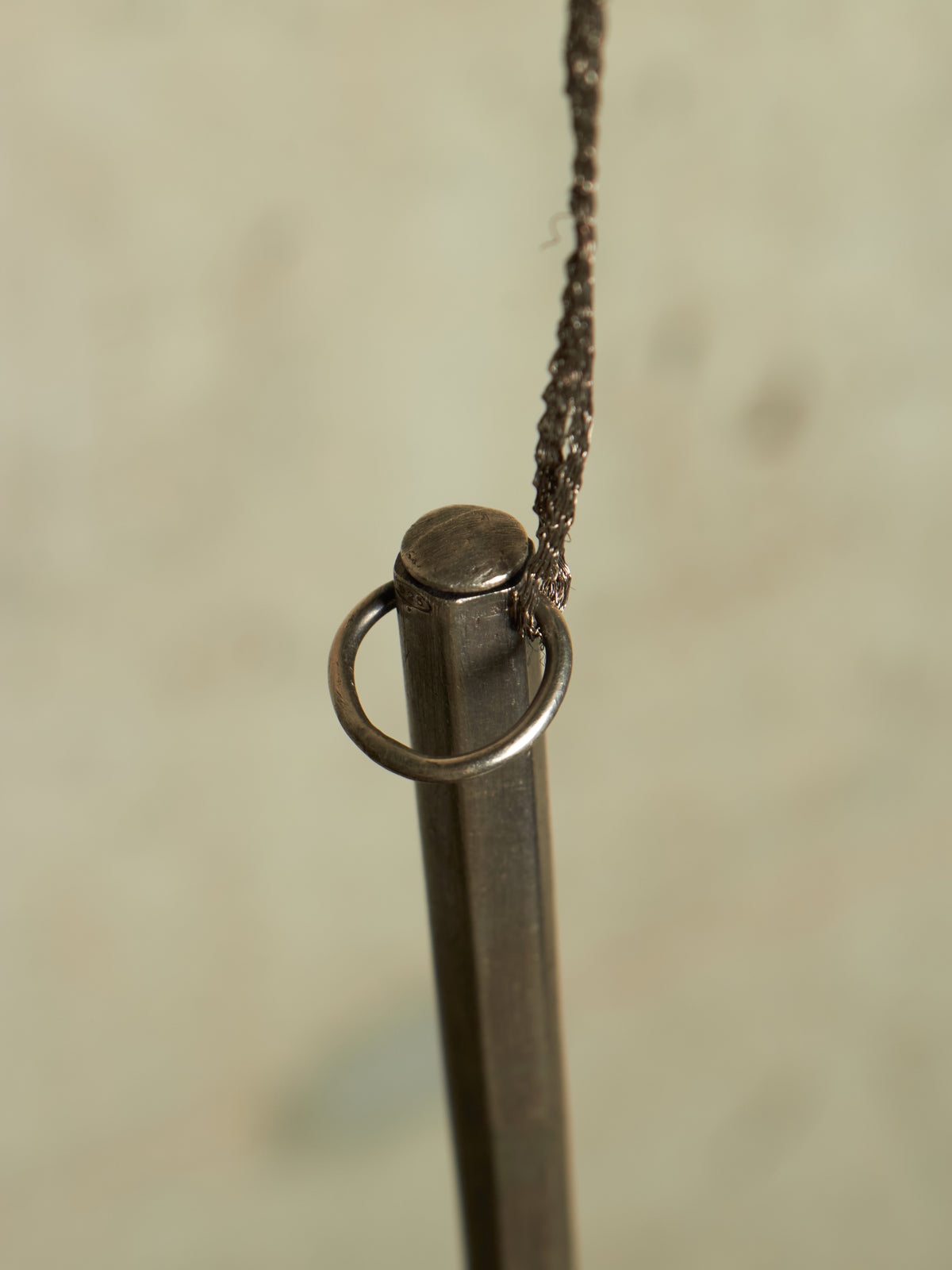 A close-up of a dark metal hanging rod with a circular ring near the top and a thin dark cord attached, set against a softly blurred light beige background.