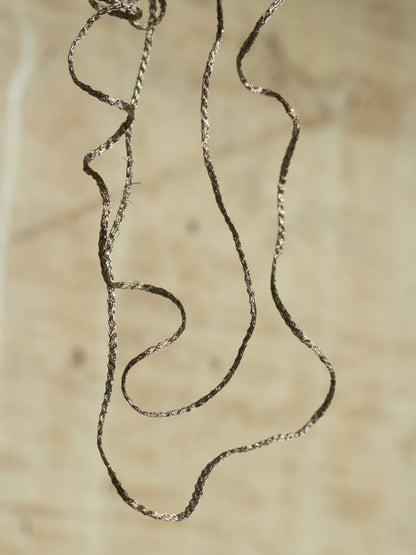A close-up of a thin, twisted metallic chain or cord in a dark silver/bronze tone, draped loosely against a warm beige textured background.