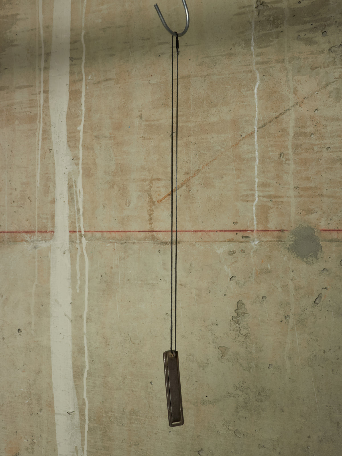 A minimalist metal wall hanging with a long black cord and a rectangular dark brown wooden weight, suspended from a metal hook against a textured concrete wall.