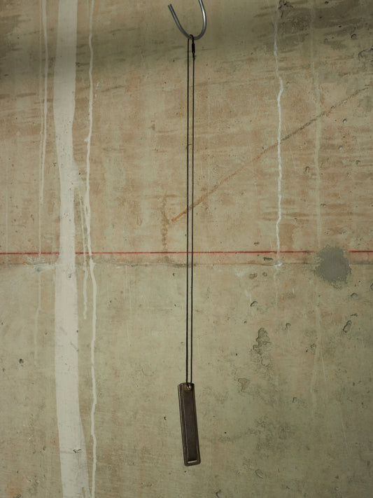 A minimalist metal wall hanging with a long black cord and a rectangular dark brown wooden weight, suspended from a metal hook against a textured concrete wall.