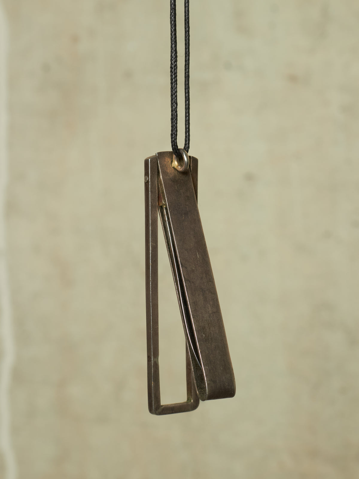 A minimalist, geometric open-frame pendant in a dark, antique bronze or oxidized metal finish, suspended from a black cord against a textured, warm beige background.