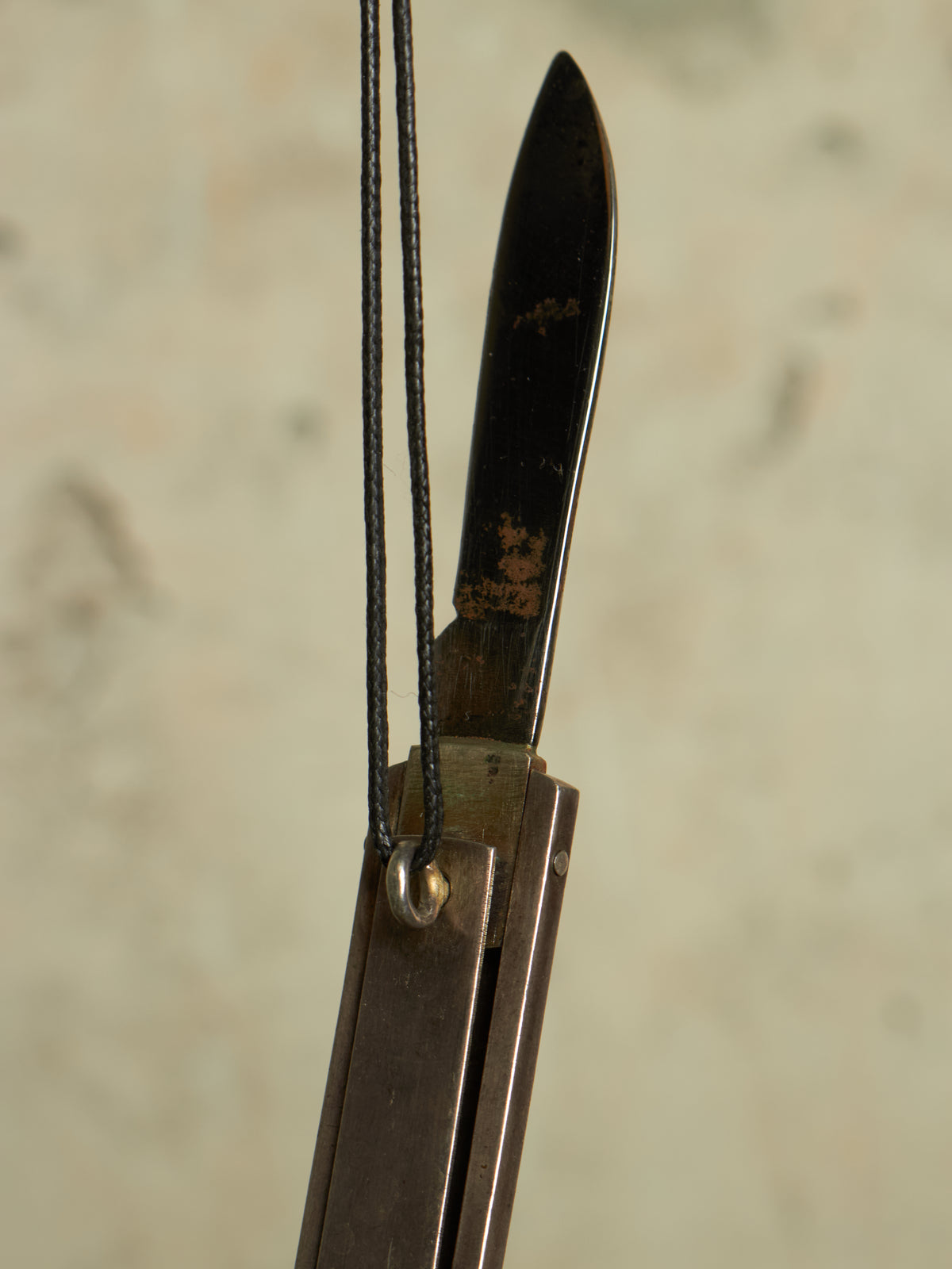 A close-up of a folding bow component, likely a riser or stabilizer, featuring a dark metal blade with a weathered patina and a black cord loop attached near the center.