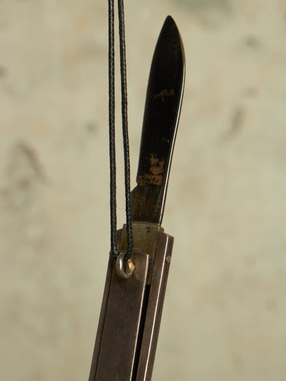 A close-up of a folding bow component, likely a riser or stabilizer, featuring a dark metal blade with a weathered patina and a black cord loop attached near the center.