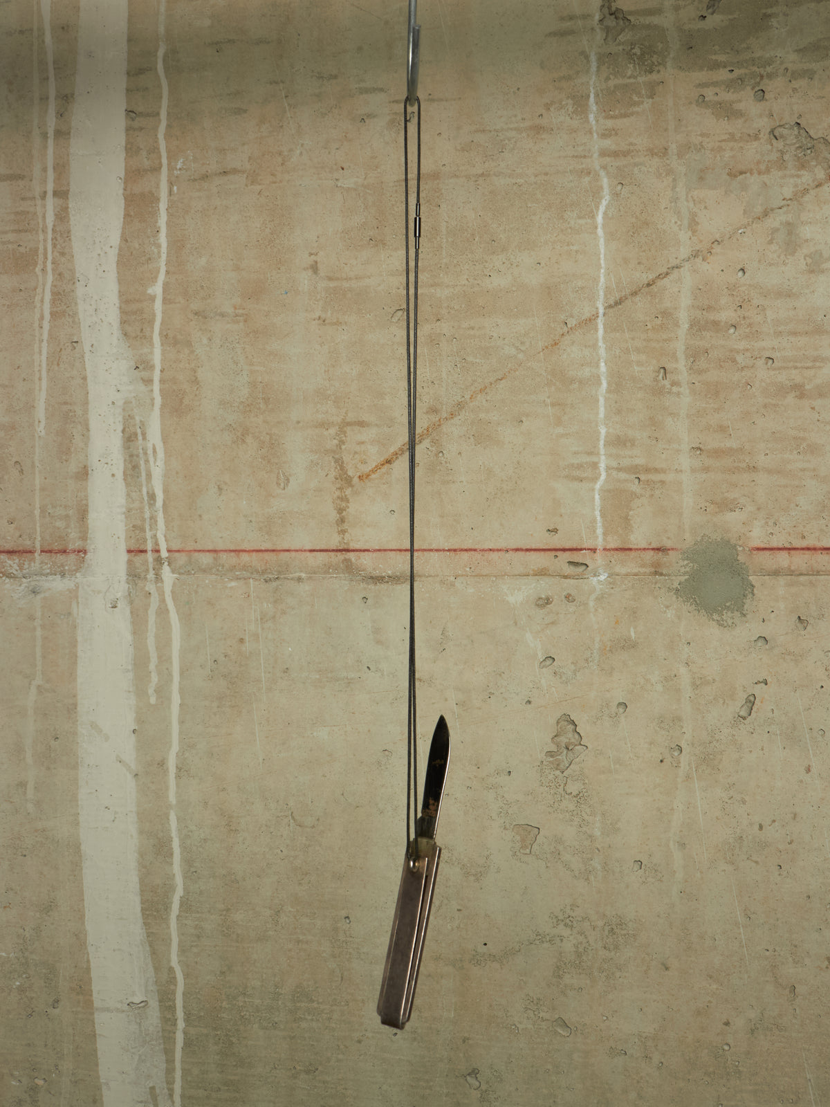 A folding pocket knife with a dark handle and black blade is suspended vertically by a thin metal rod against a textured concrete wall with white paint drips and a faint red horizontal line.