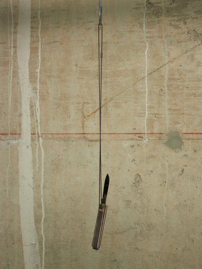 A folding pocket knife with a dark handle and black blade is suspended vertically by a thin metal rod against a textured concrete wall with white paint drips and a faint red horizontal line.
