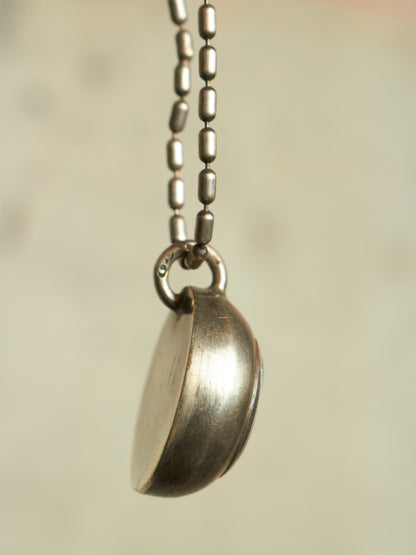 A close-up of a silver pendant necklace featuring a small, semi-circular dish or locket pendant on a matching silver bead chain, set against a soft, warm beige background.