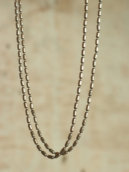 A double-strand necklace made of small metallic oval beads, shown hanging against a soft beige textured background.