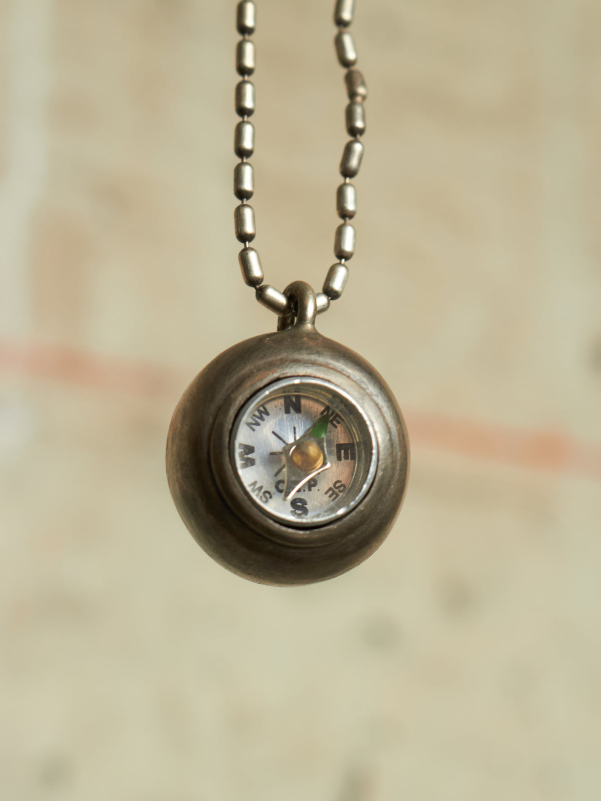 A small round compass pendant necklace with a metallic bronze finish and a ball chain, shown against a softly blurred beige background.