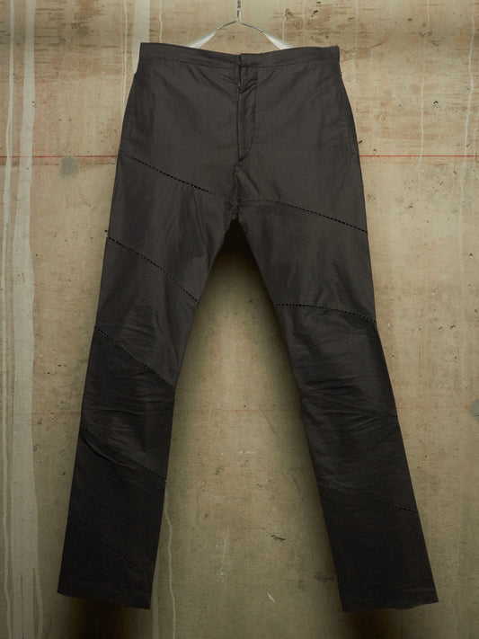 Dark charcoal gray pants with diagonal seam details, hanging on a wire hanger against a textured concrete wall.