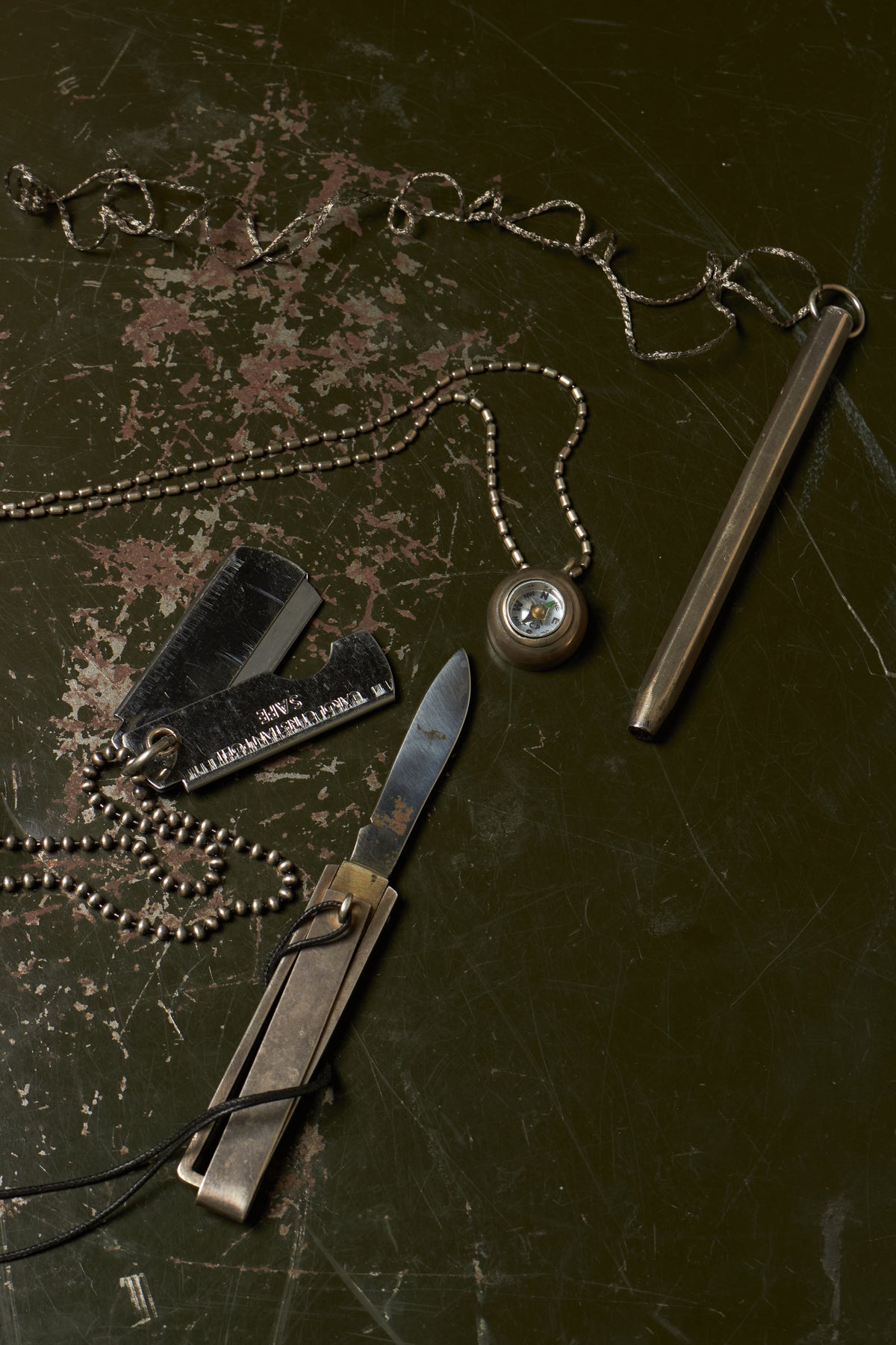 A survival and outdoor gear set arranged on a dark, distressed surface, including a metal fire starter rod, a folding knife, dog tags, a compass, and a beaded chain necklace.