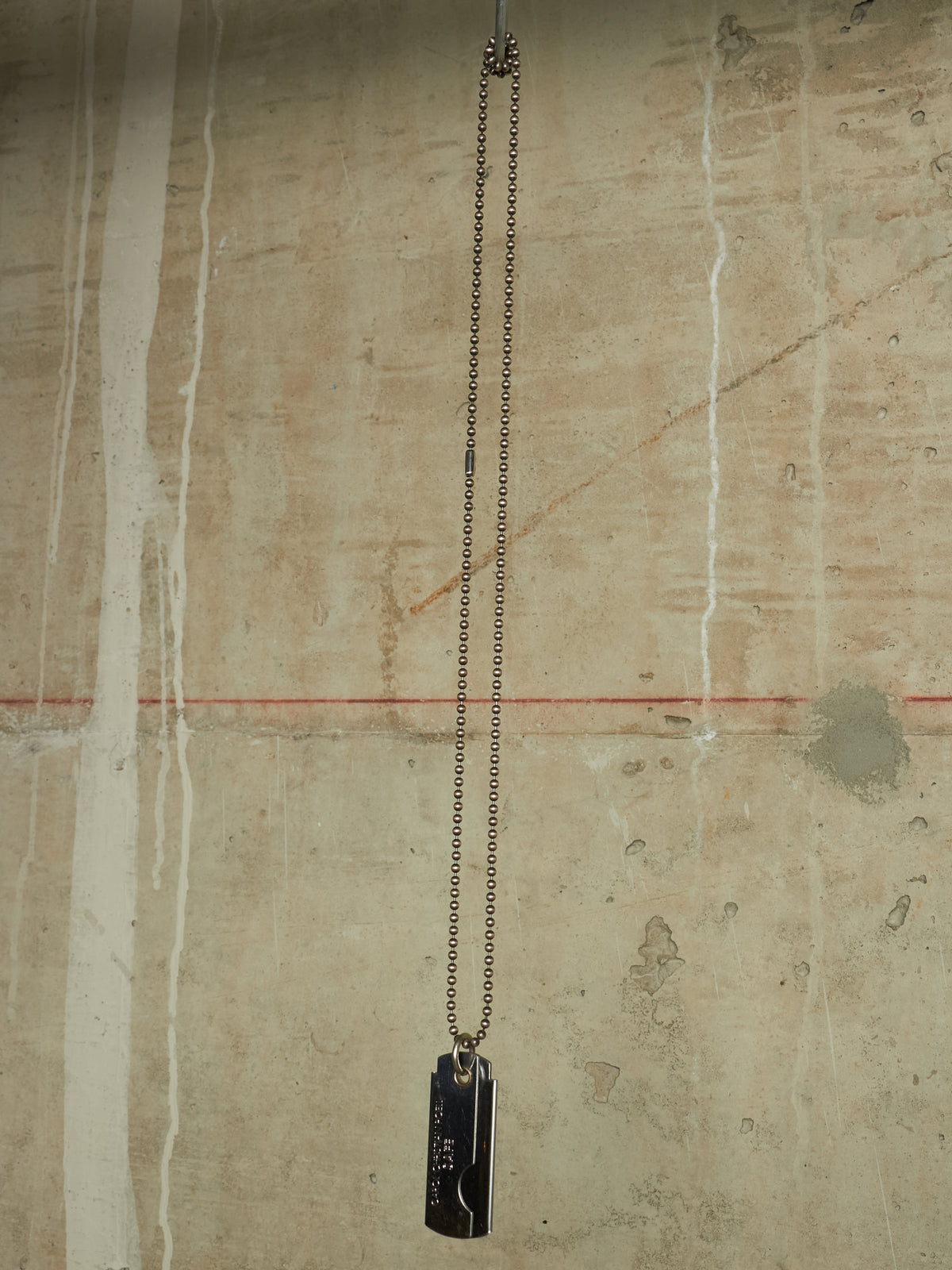 A black metal dog tag pendant necklace hanging from a silver-toned chain against a textured concrete wall.